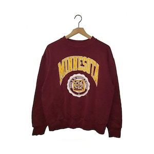 Könnte beinhalten: Ein kastanienbraunes Sweatshirt mit Rundhalsausschnitt, das den Schriftzug MINNESOTA in großen, gelben, gebogenen Buchstaben über einem kreisförmigen Siegel mit der Aufschrift REGENTS OF THE UNIVERSITY OF MINNESOTA zeigt. Gerippte Bündchen.