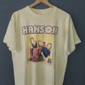 May include: Cream-colored vintage t-shirt featuring the band name "HANSON" in purple lettering above a rectangular image. The image shows three people, with a signature below. The shirt is hanging on a wooden hanger.