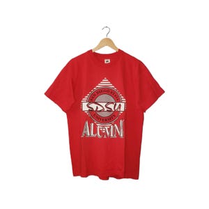 Peut inclure: T-shirt rouge avec un motif graphique. Le motif comprend le texte "SDSU UNIVERSITY ALUMNI" en blanc et gris. Le graphisme comporte également les mots "SAN DIEGO STATE" dans un cercle. Le t-shirt est suspendu à un cintre en bois.
