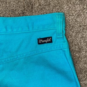 May include: A pair of turquoise blue denim jeans with a Wrangler logo on the back pocket.
