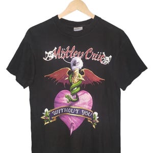 以下が含まれることがあります： 黒いTシャツ。バンド名「Mötley Crüe」が白い文字で書かれています。グラフィックには、翼のある剣、緑色のヘビ、紫色のハートがあり、「Without You」というテキストがバナーに表示されています。