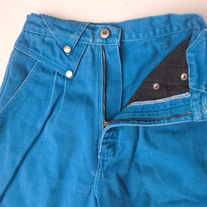 May include: Close-up of a pair of bright blue denim pants. The pants have a zipper fly, a button closure, and two decorative buttons on the front. The pockets are visible, and the pants appear to be high-waisted.