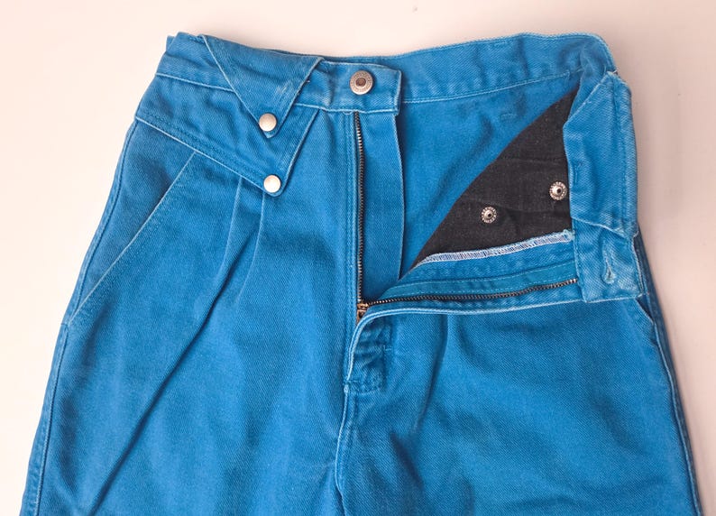 May include: Close-up of a pair of bright blue denim pants. The pants have a zipper fly, a button closure, and two decorative buttons on the front. The pockets are visible, and the pants appear to be high-waisted.