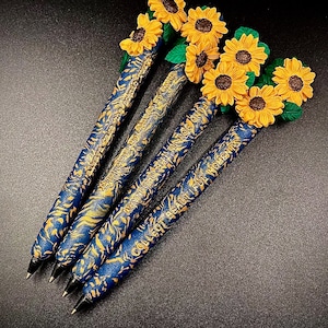 May include: Five blue and gold pens with sunflower toppers. The pens have a repeating pattern of gold swirls on a blue background. The text on the pens reads "Collect Beauty, Find Moments".
