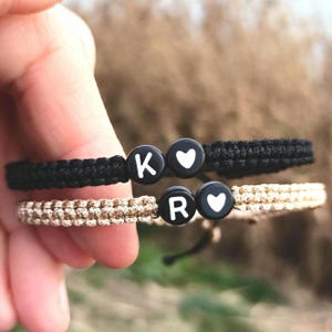 May include: Two friendship bracelets with black and beige cords. The bracelets have black beads with white letters and hearts. The letters on the beads spell "K" and "R" with a heart on each bracelet.
