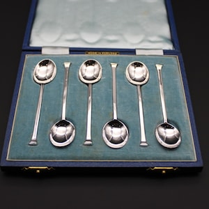 Puede incluir: Un juego de seis cucharas de plata presentadas en una caja forrada de terciopelo azul. Las cucharas tienen cuencos redondeados y mangos largos y delgados. La caja rectangular tiene una tapa con bisagras y cierres dorados.