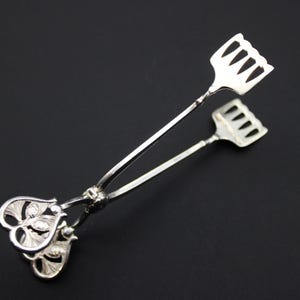 May include: Silver-toned serving tongs with a decorative floral design on the handle and a fork-like end. The tongs are made of a shiny metal and are set against a dark background.