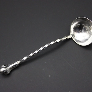 Puede incluir: Una cuchara de plata con mango retorcido y cuenco redondeado. El mango tiene un extremo decorativo y bulboso. La cuchara está hecha de un metal brillante y reflectante y está sobre un fondo oscuro.