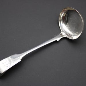 May include: A silver ladle with a rounded bowl and a long, slender handle. The handle has a decorative design near the bowl and a monogram at the end. The ladle is made of a shiny, reflective metal.