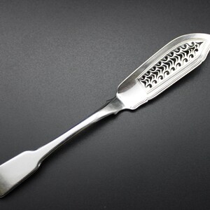 Puede incluir: Un cuchillo de mantequilla de plata con un diseño decorativo calado en la hoja. El mango es liso y redondeado.