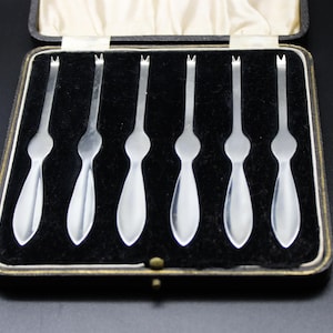 May include: A set of six silver cocktail forks in a black velvet lined box. The forks are arranged in a row and have a pointed tip and a rounded handle.