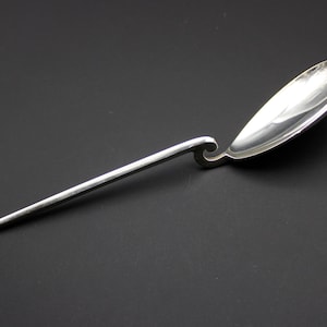 May include: A silver spoon with a long, slender handle and a teardrop-shaped bowl. The handle has a decorative swirl near the bowl. The spoon is set against a dark background.