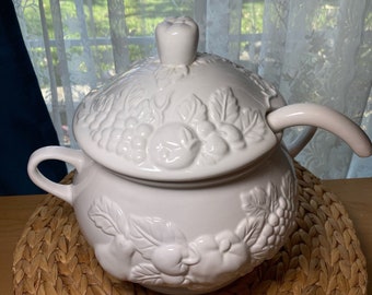 May include: White ceramic soup tureen with a lid and handle. The tureen is decorated with embossed fruit and leaf designs. The lid is topped with a small apple-shaped knob. The tureen sits on a woven placemat.