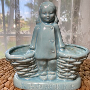 May include: A light blue ceramic figurine of a girl holding two woven baskets. The girl wears a coat with two buttons. The base of the figurine has Chinese characters. The figurine is on a woven mat.