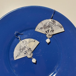 Op de afbeelding: Een paar zilverkleurige, waaiervormige oorbellen met ingewikkelde ontwerpen. Elke oorbel heeft een kleine, witte, ronde kraal die aan de onderkant hangt. De oorbellen worden gepresenteerd op een blauw bord.