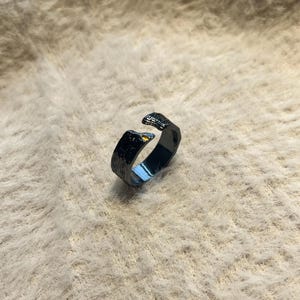 Op de afbeelding: Een donkergrijze ring met een getextureerd oppervlak. De ring heeft een open ontwerp met twee verhoogde, getextureerde uiteinden. De binnenkant van de ring reflecteert een gouden tint. De ring is een sieraad.