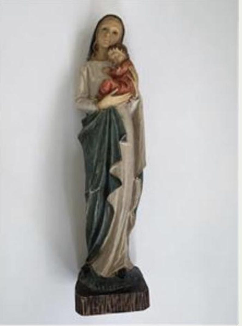 May include: A carved wooden statue of a woman holding a child. The woman wears a long, flowing beige robe with a dark green drape. The child is dressed in red. The statue is mounted on a dark brown base.