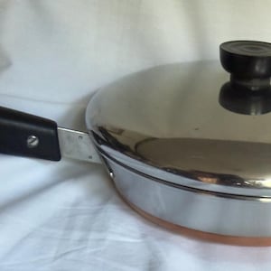 Op de afbeelding: Een roestvrijstalen koekenpan met een koperkleurige bodem en een bijpassend deksel. De pan heeft een lange, zwarte, hittebestendige handgreep met een metalen ophanglus aan het uiteinde. Het deksel heeft een centrale, zwarte knop.