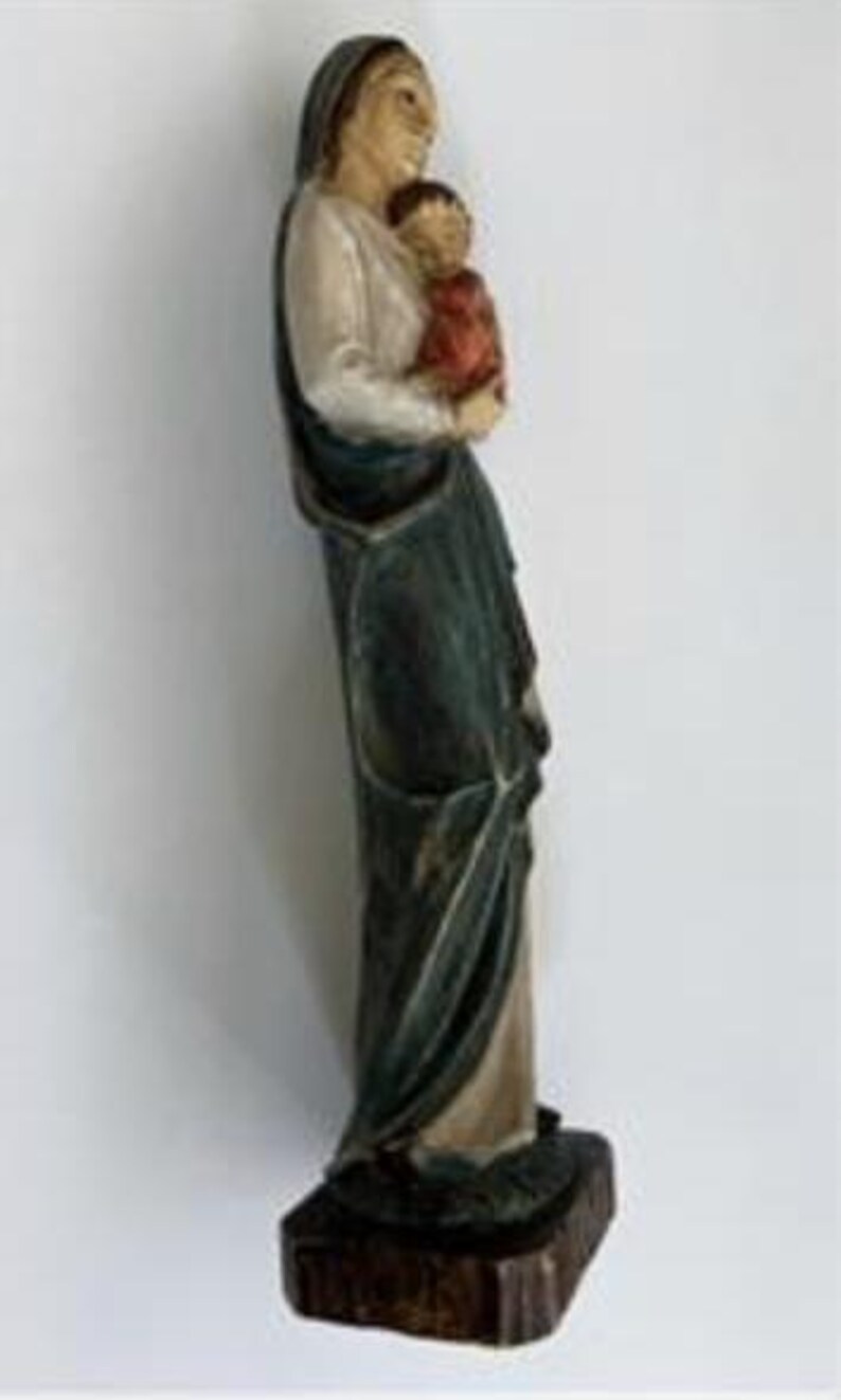 May include: A carved wooden statue of a woman holding a child. The woman wears a white top, a dark green cloak, and a head covering. The child is dressed in red. The statue is mounted on a dark brown wooden base.