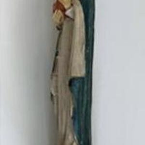 May include: A carved wooden statue of a religious figure, featuring a person in a long robe and a dark blue cloak, holding a red object. The statue has a weathered appearance and stands on a dark, textured base.