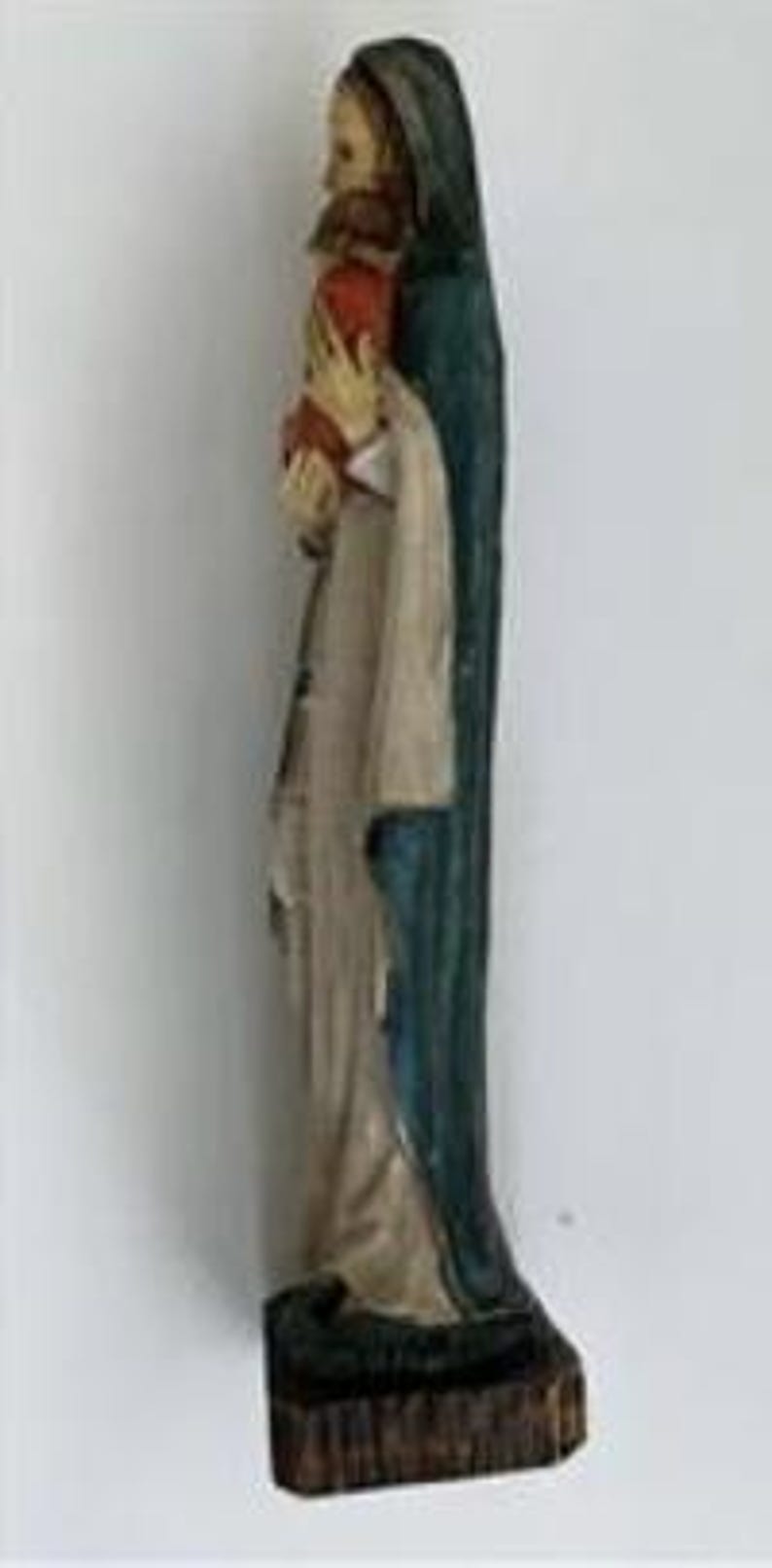 May include: A carved wooden statue of a religious figure, featuring a person in a long robe and a dark blue cloak, holding a red object. The statue has a weathered appearance and stands on a dark, textured base.
