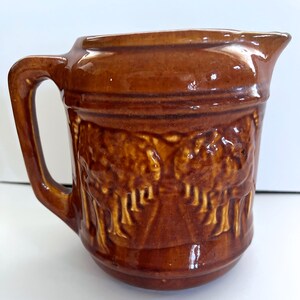 May include: A brown ceramic pitcher with a handle and spout. The pitcher features a detailed embossed design of trees or foliage around the body. The glaze gives it a glossy finish, and the overall style suggests a vintage or rustic aesthetic.