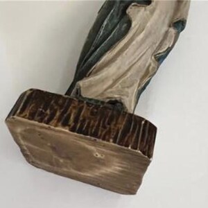 May include: A religious figurine with a rectangular base. The base is brown with a textured, wood-like appearance. The figurine's clothing is off-white and blue, with a detailed, sculpted design.