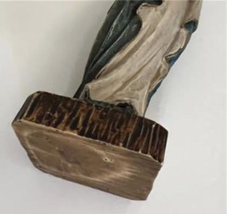 May include: A religious figurine with a rectangular base. The base is brown with a textured, wood-like appearance. The figurine's clothing is off-white and blue, with a detailed, sculpted design.