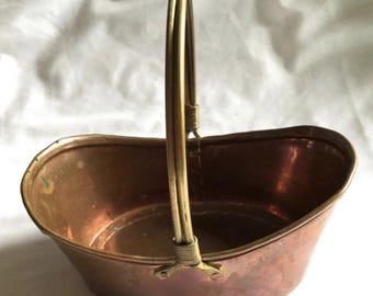 May include: An oval-shaped copper basket with a polished brass handle. The basket features a smooth, metallic copper body with a warm, reddish-brown tone. The handle is constructed from two thin brass rods, secured at the base with wire wrapping.