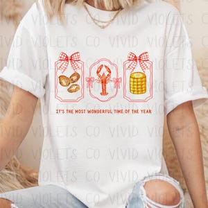 May include: White t-shirt with a red, white, and blue design. The design features three framed images with a bow on top of each frame. The images are of crawfish, corn on the cob, and potatoes. The text "It's the most wonderful time of the year" is printed below the images.