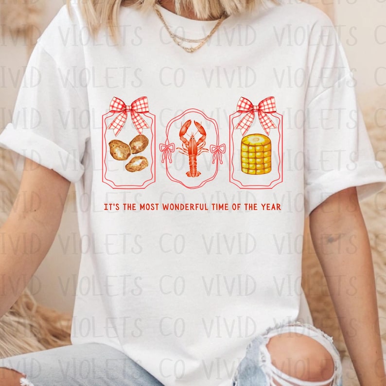 May include: White t-shirt with a red, white, and blue design. The design features three framed images with a bow on top of each frame. The images are of crawfish, corn on the cob, and potatoes. The text "It's the most wonderful time of the year" is printed below the images.