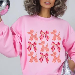 May include: Pink sweatshirt with a graphic print of pink and red balloon dogs and bows.