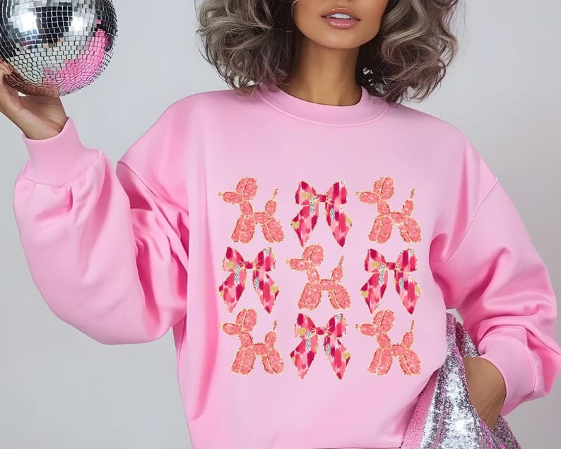 May include: Pink sweatshirt with a graphic print of pink and red balloon dogs and bows.