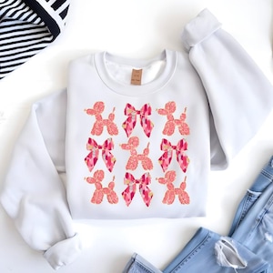 May include: White sweatshirt with a pink and orange graphic print of multiple balloon dogs with bows.