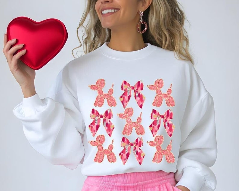 May include: White sweatshirt with a pink and white graphic print of multiple balloon dog and bow designs.