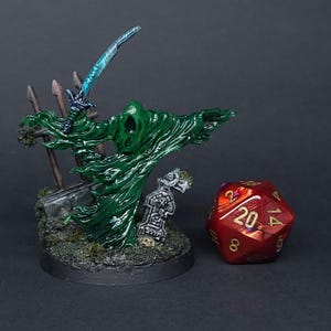 May include: A green and blue painted miniature figure of a ghost holding a sword, standing on a base with a graveyard theme. The figure is next to a red 20-sided die with gold numbers.
