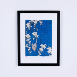 May include: Framed art print featuring a Japanese-style illustration. The artwork depicts a bird perched on a flowering branch against a blue background. The print is enclosed in a black frame, ready for display. The image includes Japanese characters.