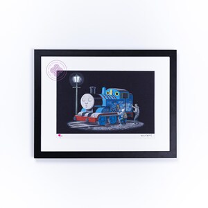 May include: A framed art print featuring a blue steam locomotive with a face, covered in graffiti, standing on tracks next to a street lamp at night. Two figures are shown applying paint to the side of the train. The print has a white mat.