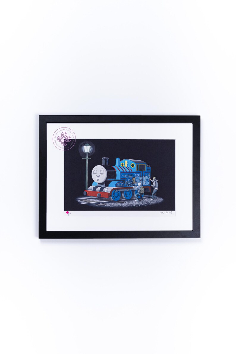 May include: A framed art print featuring a blue steam locomotive with a face, covered in graffiti, standing on tracks next to a street lamp at night. Two figures are shown applying paint to the side of the train. The print has a white mat.