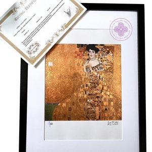 May include: A framed print of a painting by Gustav Klimt, featuring a woman in a gold dress with a mosaic pattern. The painting is titled "Adele Bloch-Bauer" and is framed in a black frame.