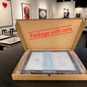 Puede incluir: Una caja de env&iacute;o de cart&oacute;n marr&oacute;n con el texto "Package with care" impreso en rojo en la tapa interior. Dentro de la caja, un objeto rectangular est&aacute; envuelto en pl&aacute;stico de burbujas transparente para proteger el contenido durante el transporte.