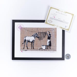 May include: A framed print featuring a black and white stencil-style illustration of a zebra and a person hanging laundry on a clothesline against a brick wall, accompanied by a white certificate of authenticity with gold decorative borders.