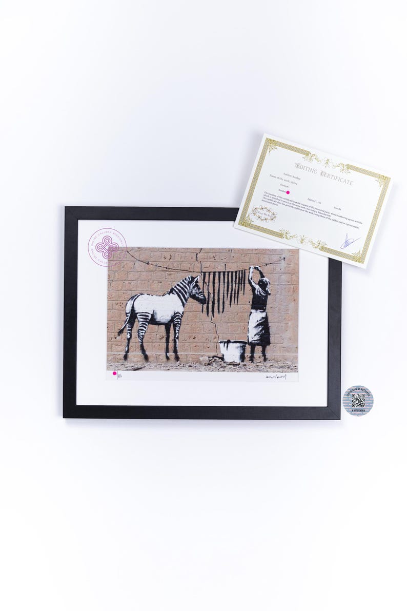 May include: A framed print featuring a black and white stencil-style illustration of a zebra and a person hanging laundry on a clothesline against a brick wall, accompanied by a white certificate of authenticity with gold decorative borders.