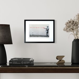 Puede incluir: Una impresi&oacute;n enmarcada en blanco y negro que muestra una figura frente a una pared, sosteniendo un cubo de pintura y un pincel. El texto "What we do in life echoes in eternity" est&aacute; escrito en rojo en la parte superior de la obra.