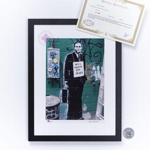 Peut inclure: Une impression encadrée montrant une personne en costume tenant une mallette devant un mur de briques vertes avec des graffitis. La personne porte un panneau indiquant WILL WORK for idiots. Un certificat d'édition à bordure dorée est posé au-dessus.
