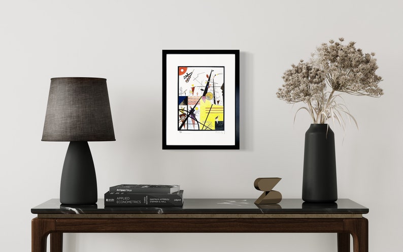 May include: A framed abstract art print with geometric shapes and lines in black, red, yellow, and white. The print is in a black frame and is hanging on a white wall.