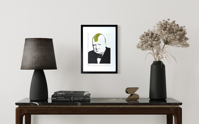 May include: A framed wall art print featuring a black and white portrait of Winston Churchill with a bright yellow mohawk hairstyle. The portrait is centered within a white mat and a thin black frame, displayed on a plain light-colored wall.