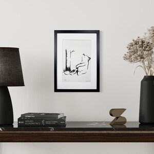 May include: A framed black and white ink illustration of a bear sitting under a tree, displayed in a black frame. The artwork is positioned on a wall above a dark console table featuring a black lamp, a stack of books, a small sculpture, and a vase.