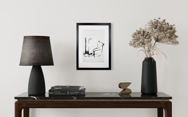 May include: A framed black and white ink illustration of a bear sitting under a tree, displayed in a black frame. The artwork is positioned on a wall above a dark console table featuring a black lamp, a stack of books, a small sculpture, and a vase.