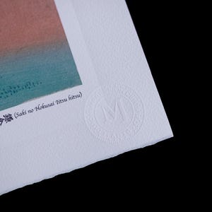 May include: A corner of a print with a landscape scene in shades of blue, green, and orange. The print has Japanese text and a circular embossed seal with the letter "M". The paper has a textured surface.